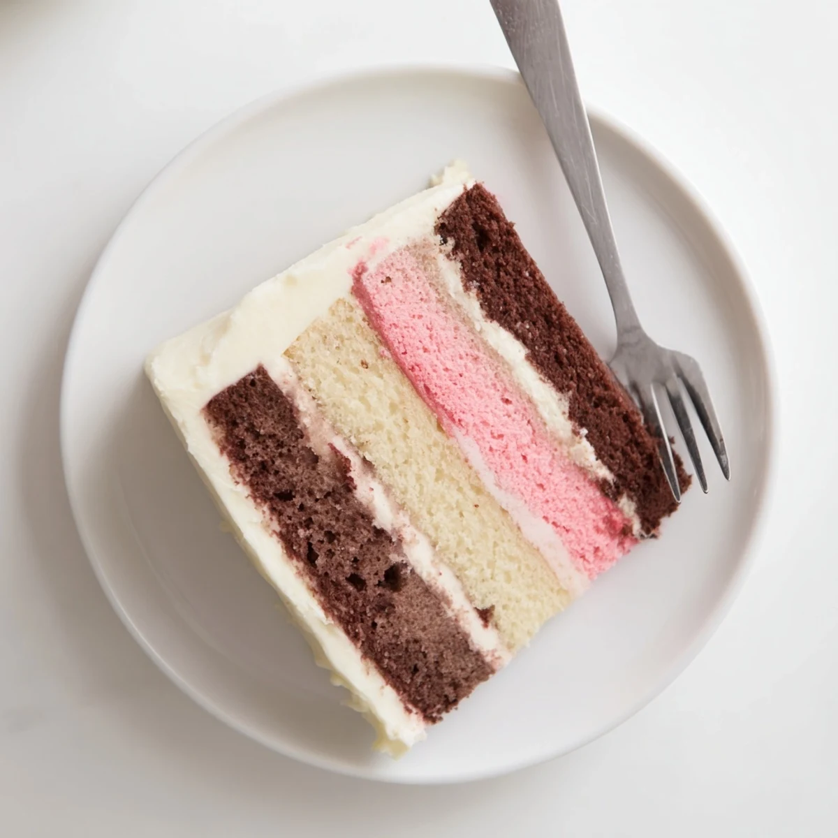 Slice of Neapolitan cake revealing three distinct colorful layers of chocolate, vanilla, and strawberry beneath creamy white buttercream frosting
