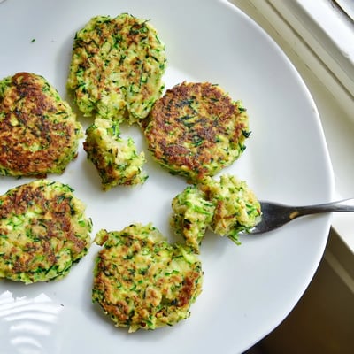 A platter of stacked Chickpea Zucchini Fritters garnished with chopped parsley, served alongside a colorful Mediterranean salad.